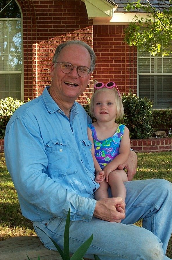 A child wearing pink sunglasses and a floral swimsuit sits with an adult in a blue shirt on a porch with brick walls.