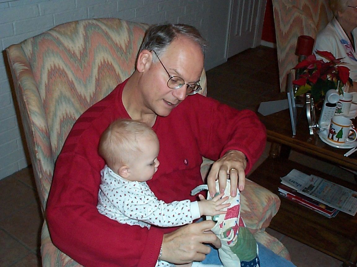 A person in a red sweater helps a small baby reach for a toy while sitting in a patterned armchair.