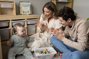 Parents sitting on floor playing and engaging with baby by clapping and playing peek-a-boo with toys.