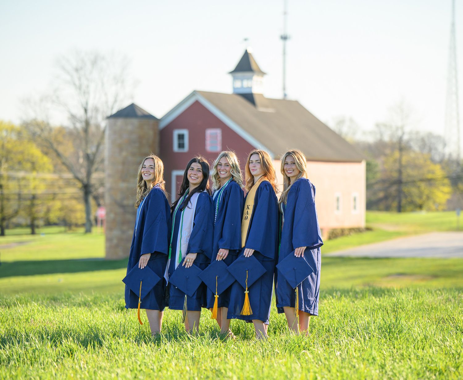 A group in blue graduation gowns and caps stand together in front of a red barn on a grassy lawn.