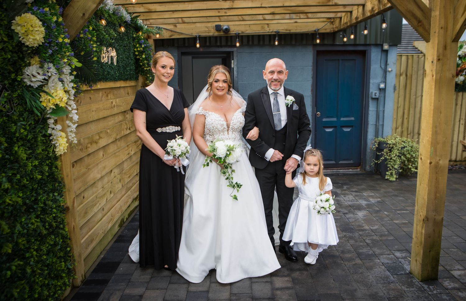 Wedding party gathered under a wooden pergola during an outdoor ceremony.