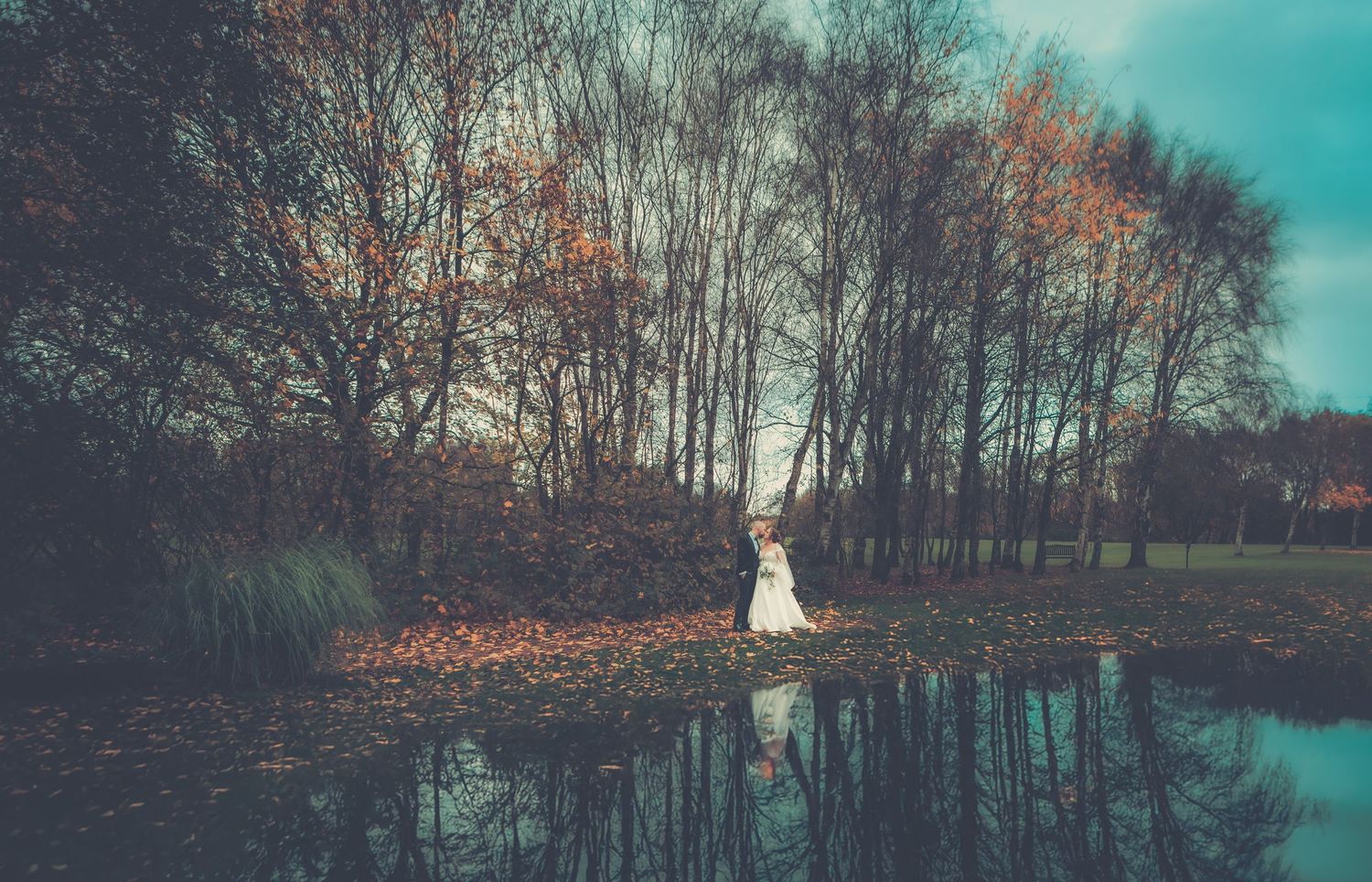 A wedding dress silhouette reflected in a pond during autumn with bare trees in the background.