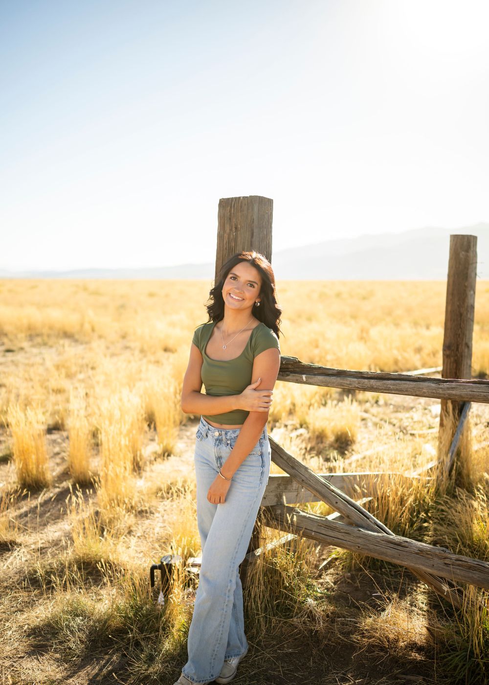 Person in green top and jeans poses near wooden fence posts in golden field.