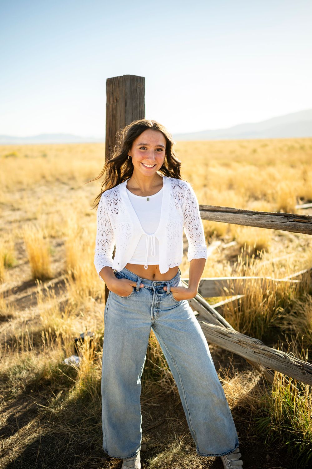 Person in white lace top and jeans poses by wooden fence in warm evening light.