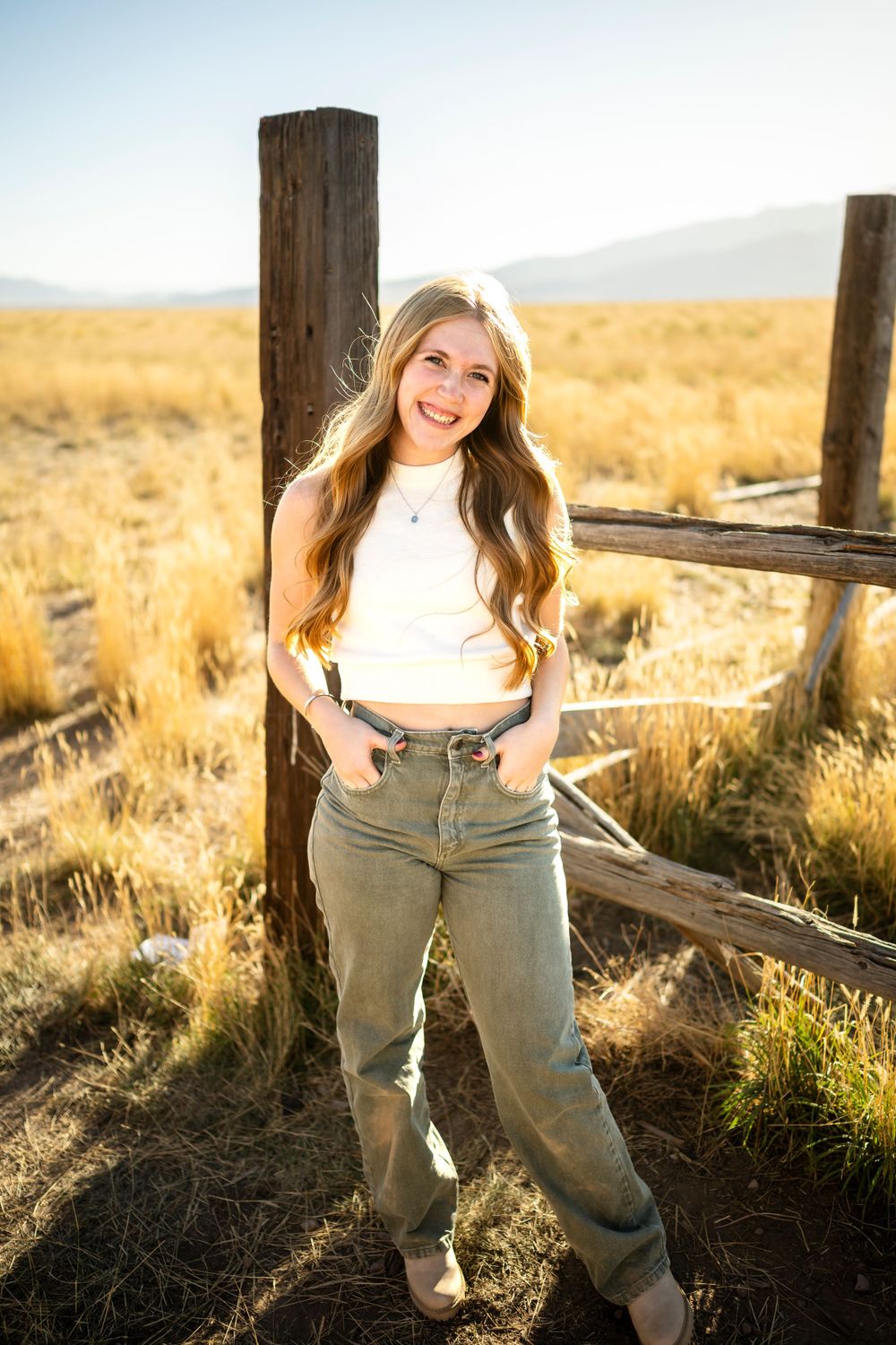 Person in white sweater and khaki pants poses playfully by rustic fence at sunset.