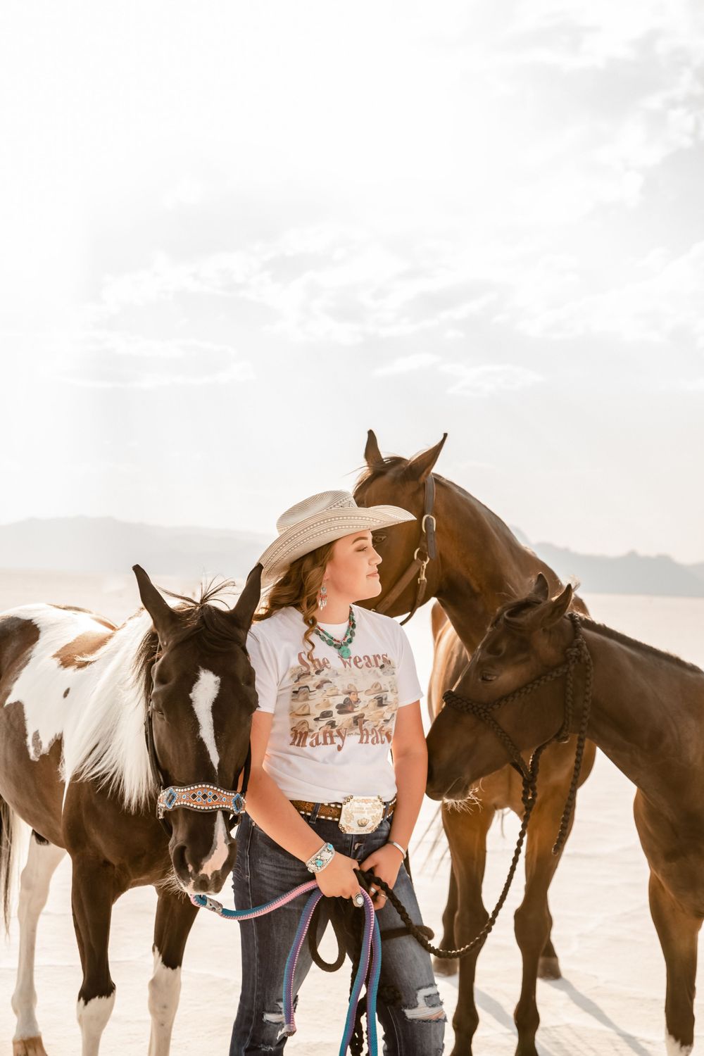 Cowgirl with three horses in desert landscape wearing western attire.