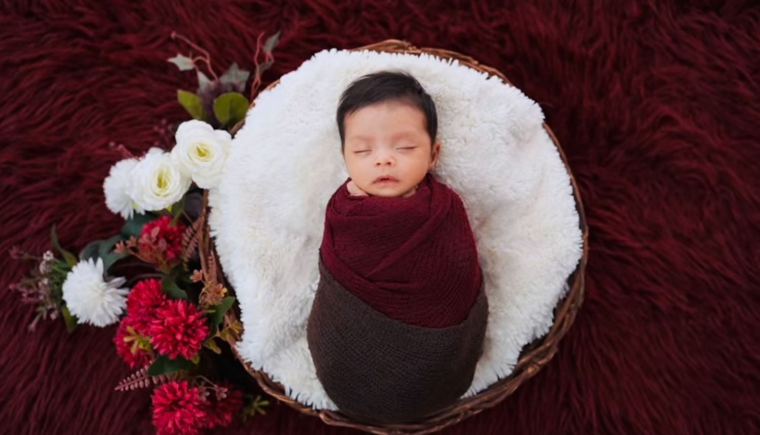 A baby portrait of a Newborn In maroon background with flowers and maroon wrapping.