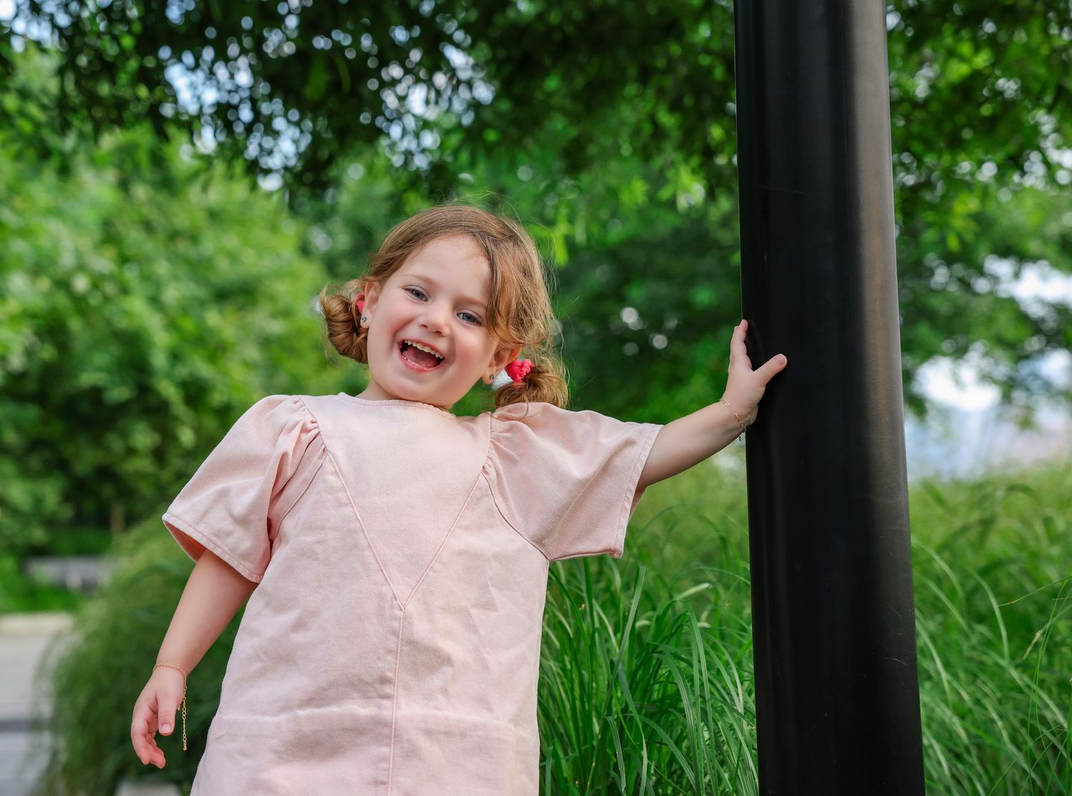 Laughing toddler girl in pink dress holding red lollipop outdoors near green trees.