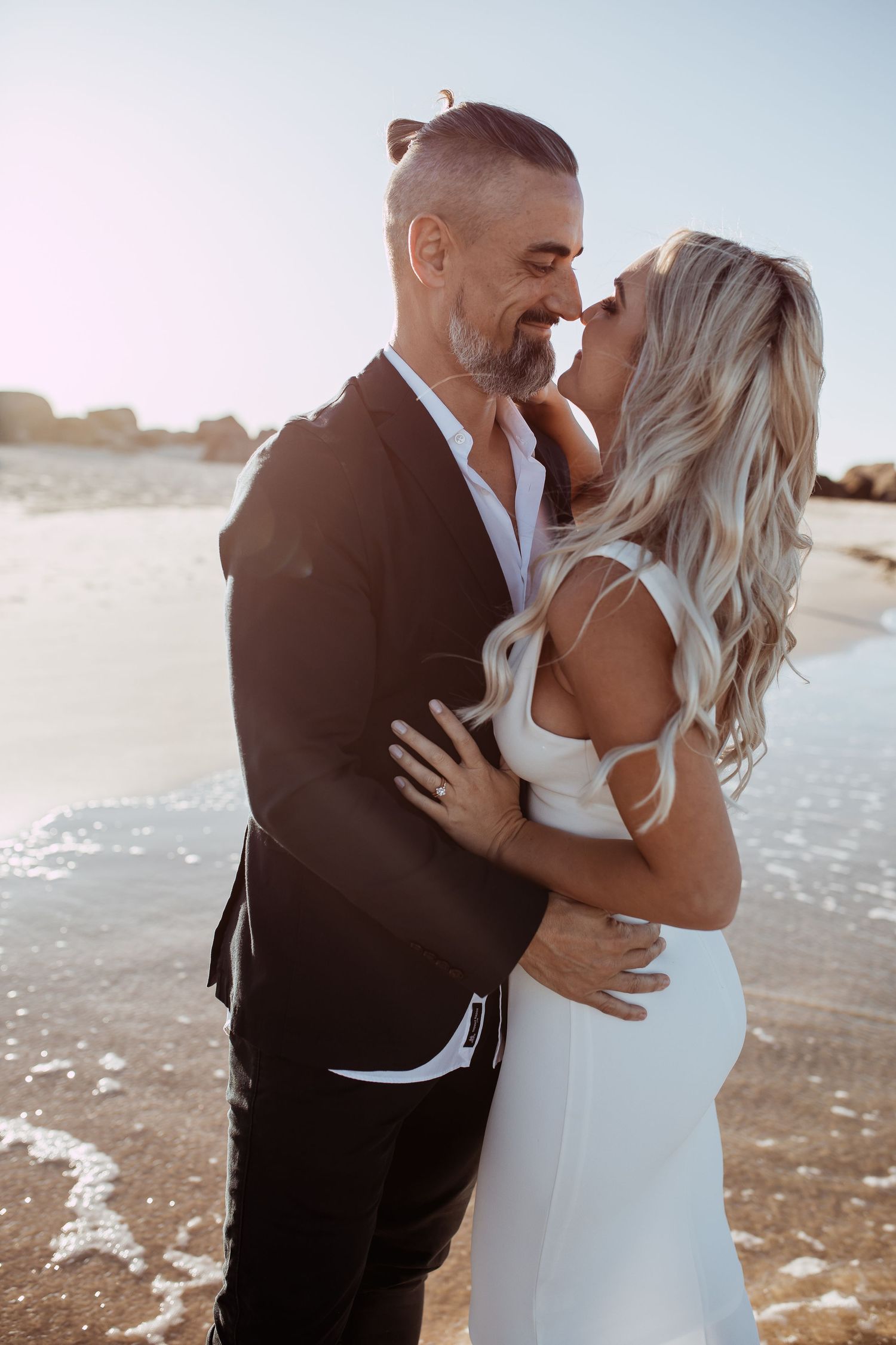A romantic couple embraces on a sunny beach, wearing formal black and white attire during a sunset photoshoot.
