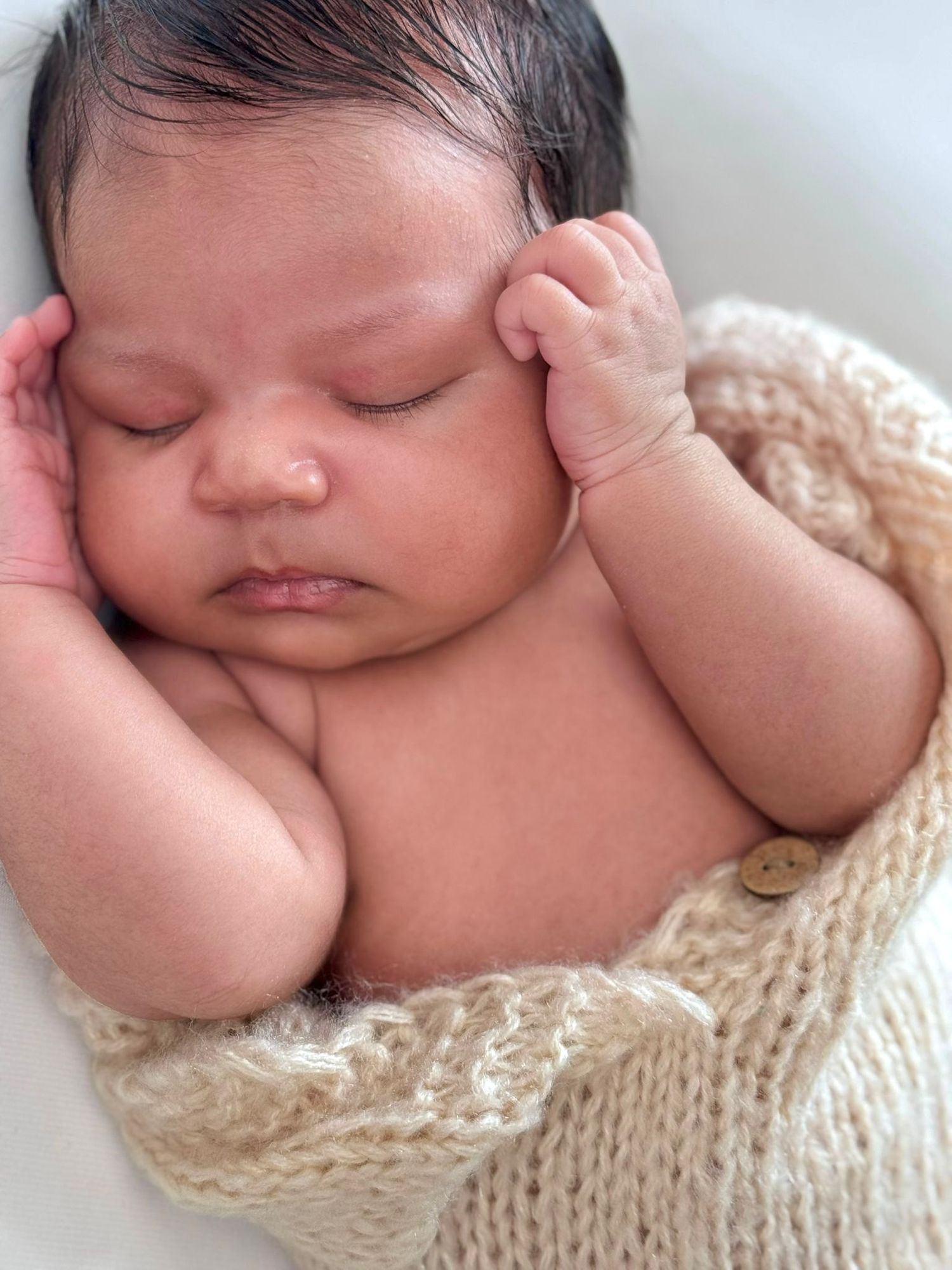 Peaceful sleeping newborn wearing soft beige knit hat rests on neutral beige backdrop in studio setting.