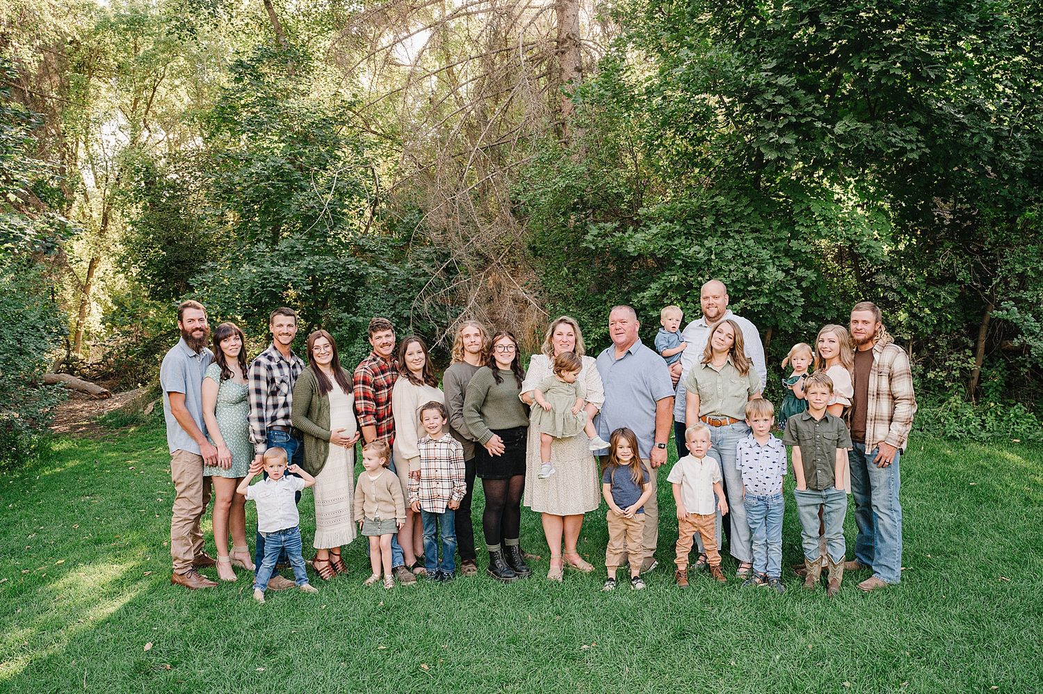Large family group portrait on a green lawn with everyone wearing coordinated casual outfits in neutral and plaid patterns.