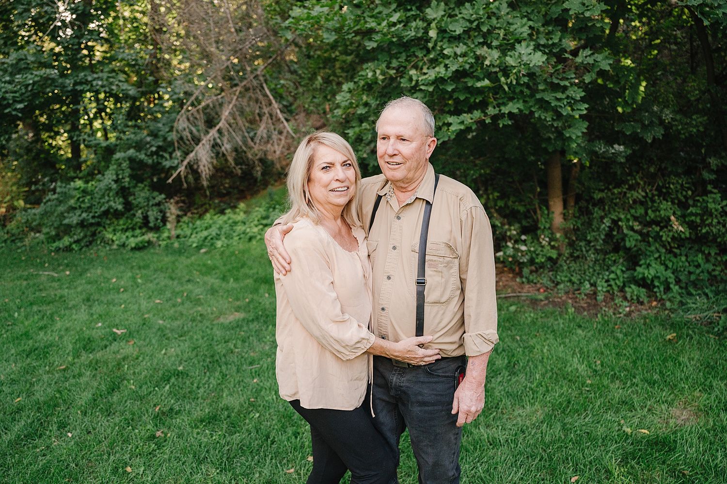 A couple stands together on a green lawn with trees and foliage in the background on a sunny day.
