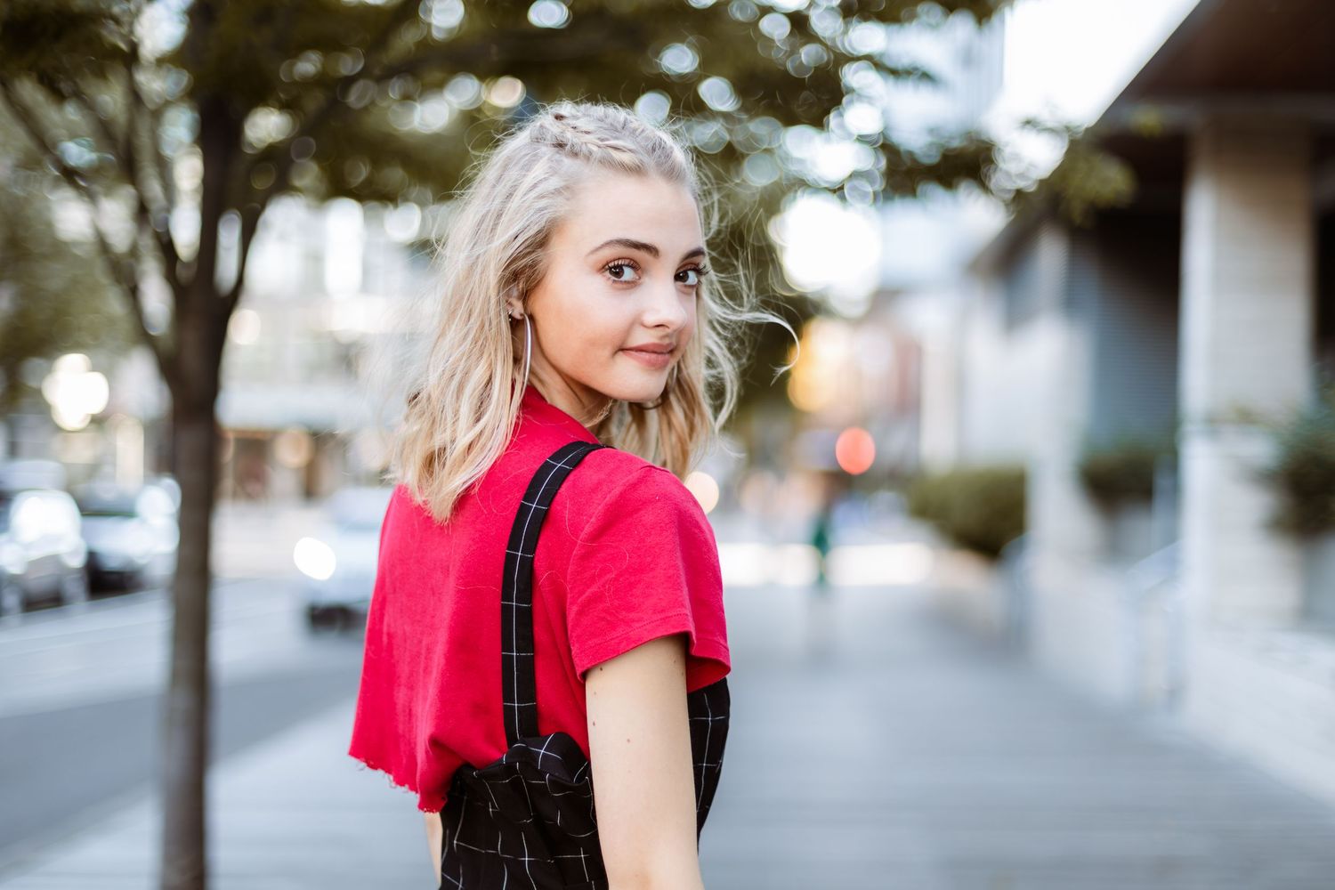 Person with blonde hair wearing a red shirt looking over their shoulder while standing on a city sidewalk.