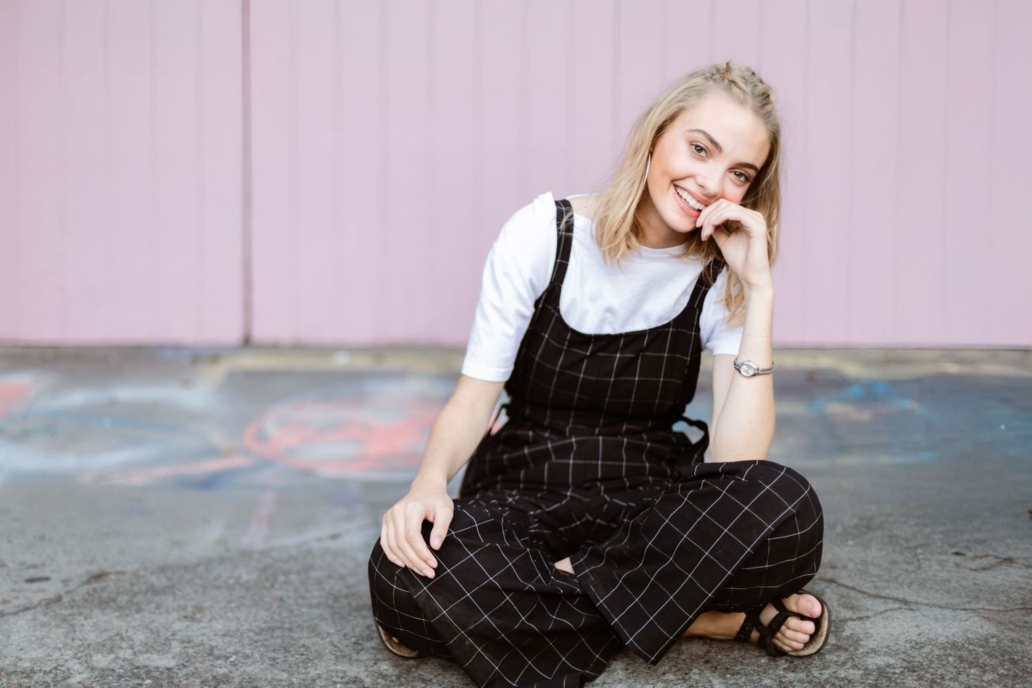 Person wearing black plaid overalls and white shirt sits cross-legged on ground against pink wall.