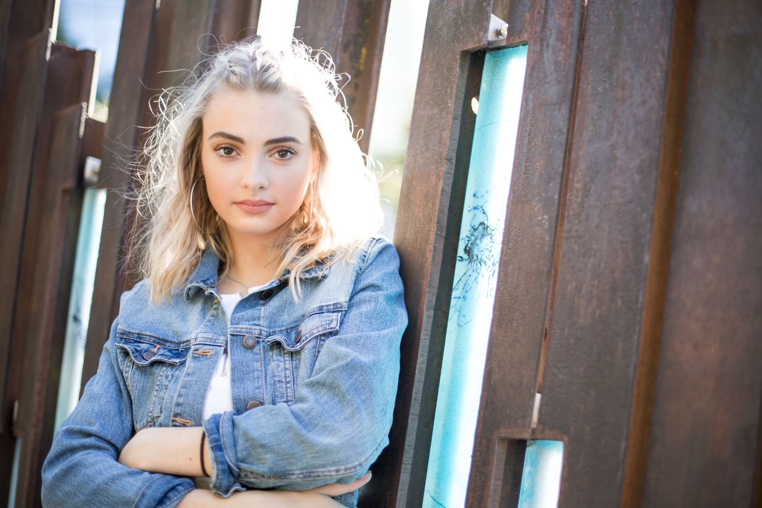 Person wearing denim jacket posing against rustic wall with arms crossed in outdoor portrait setting.