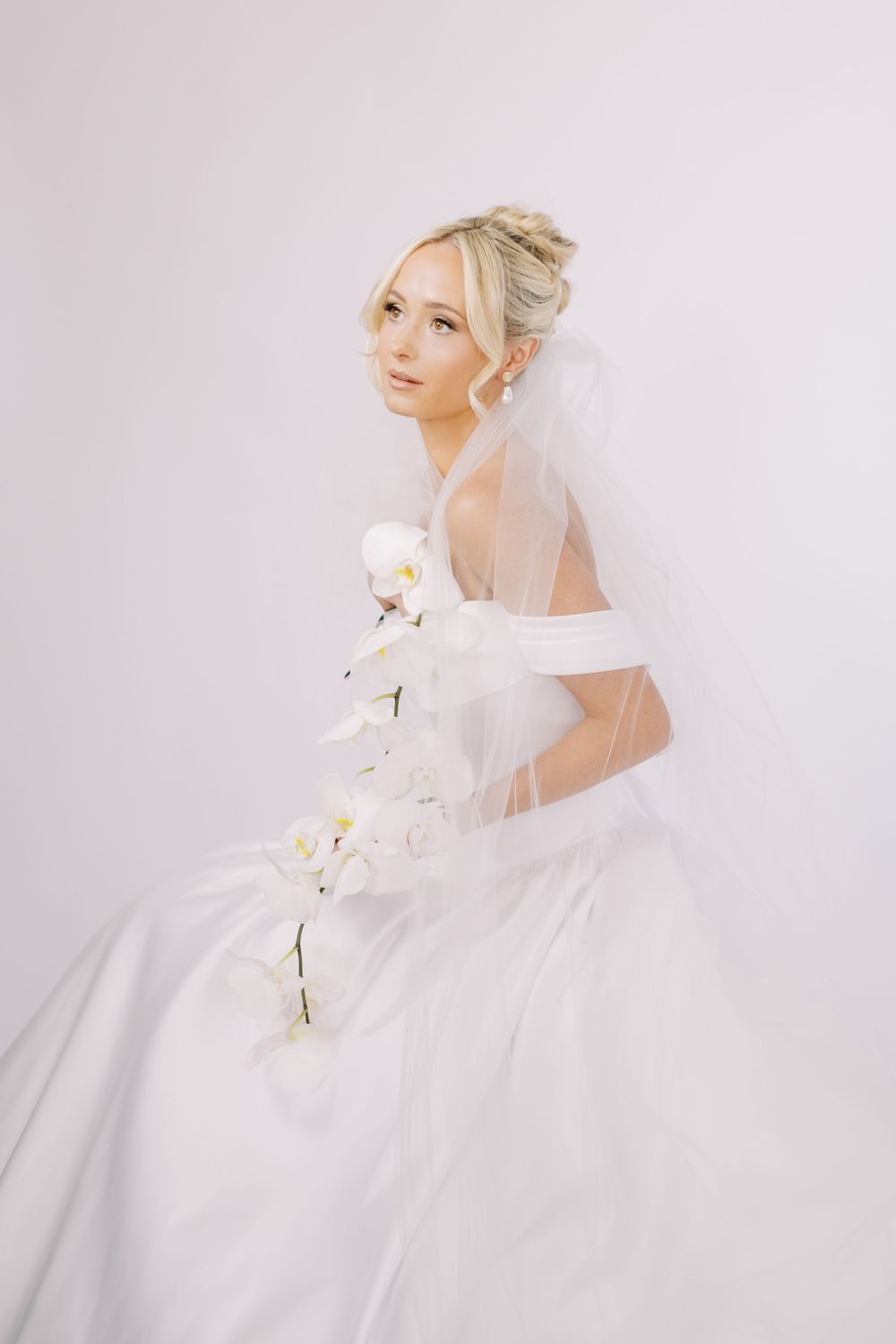 Romantic bridal portraits with flowing tulle veil and white orchids against light background.