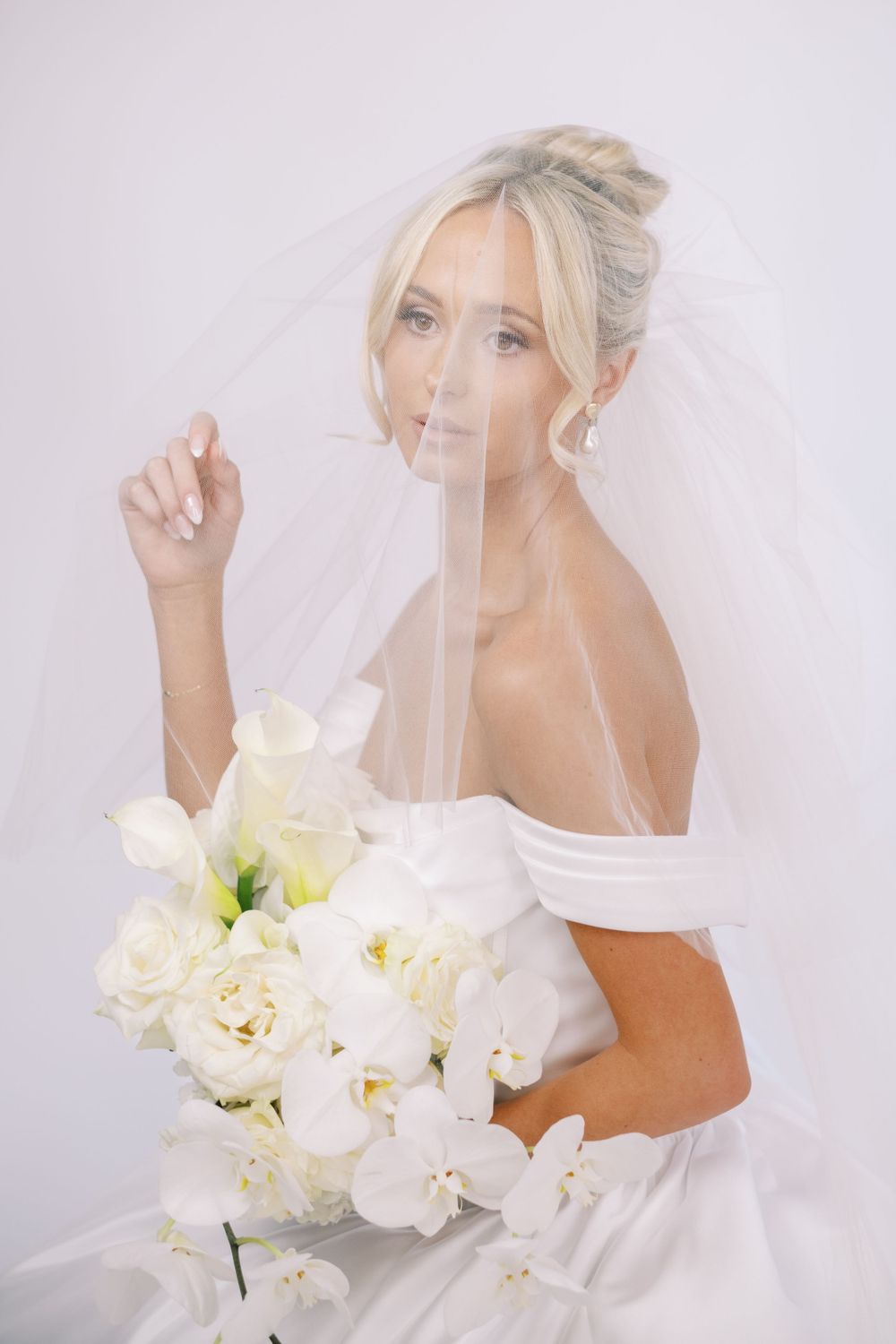 Ethereal bridal portrait with flowing veil and white orchid bouquet in bright studio setting.