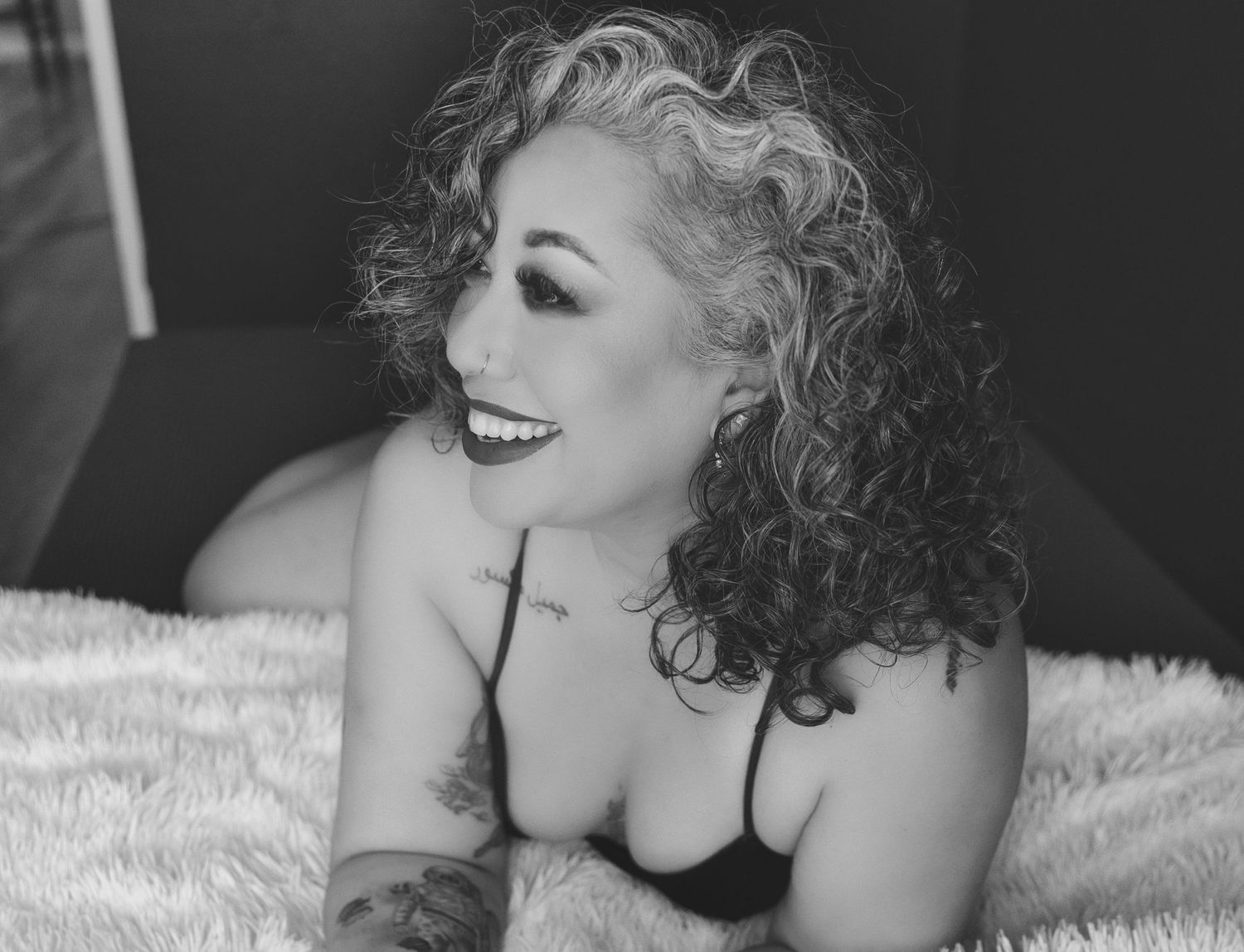 Black and white boudoir portrait of woman with curly hair smiling while lying on soft bedding.
