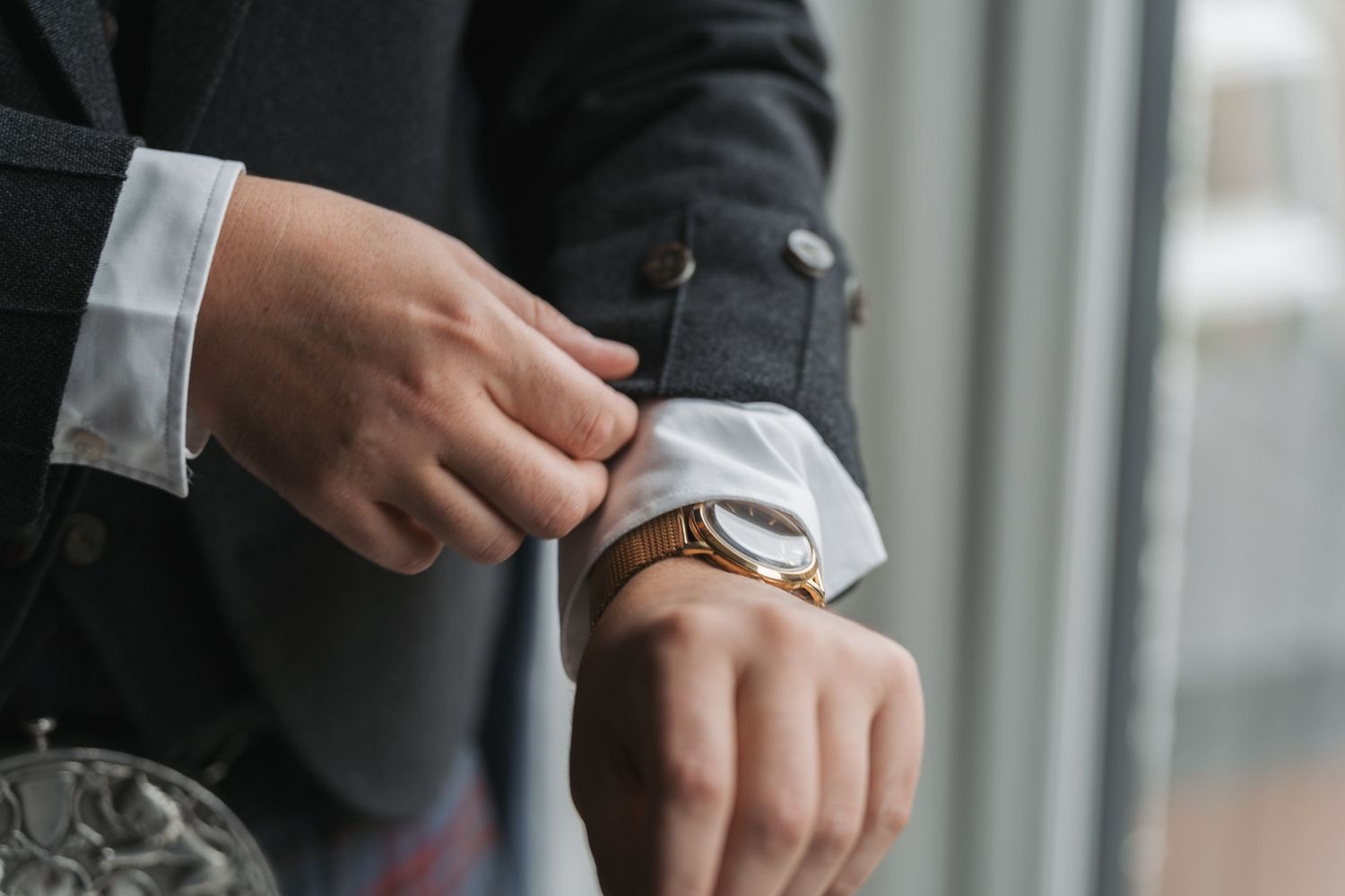 Business professional in dark suit checks elegant wristwatch while adjusting sleeve cuff near window.