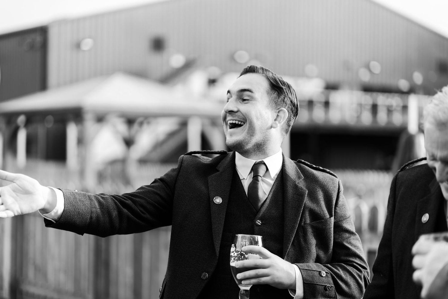 A person in a dark suit laughs joyfully while holding a drink and gesturing with their arm outstretched.