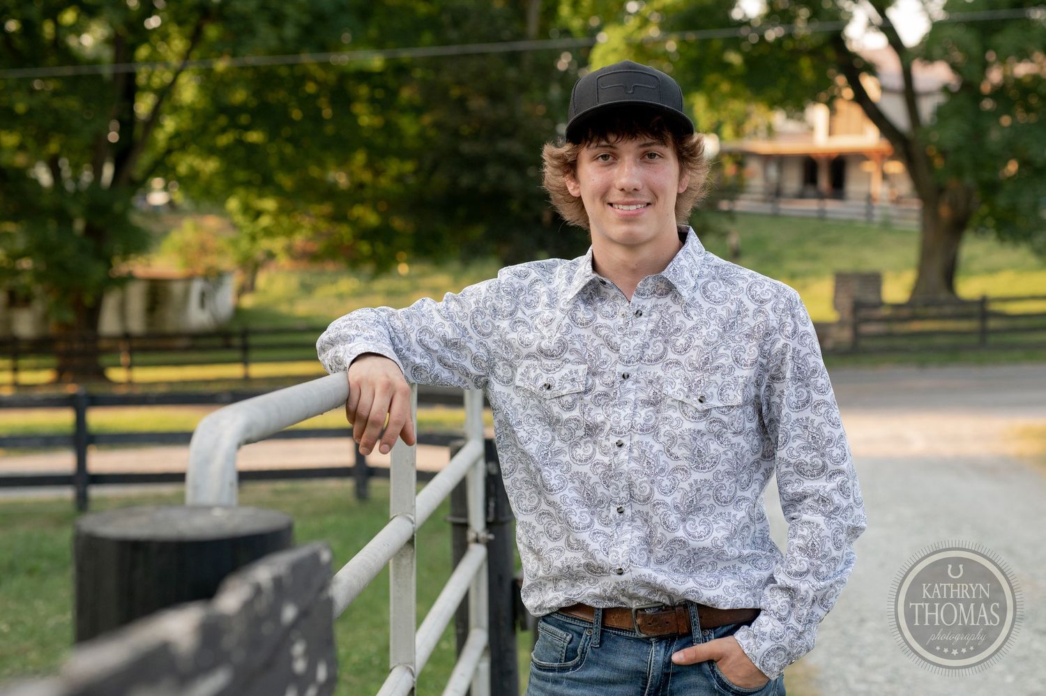 A person in a patterned shirt and baseball cap poses by a wooden fence at sunset.