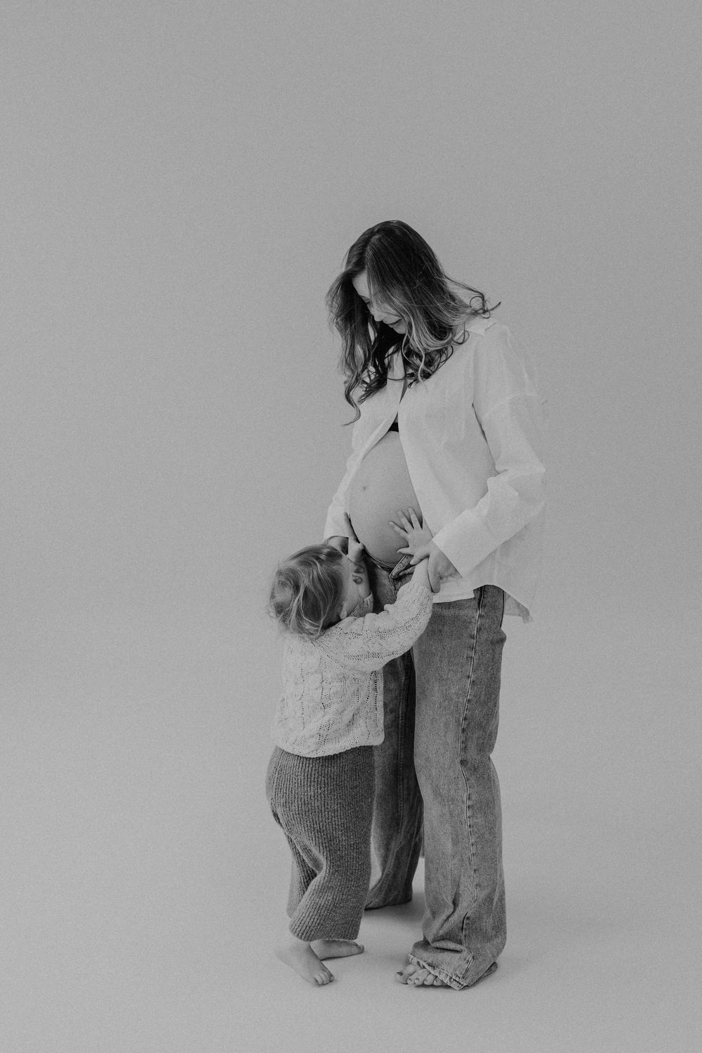 Schwarzweiß-Bild von einer Schwangeren im Fotostudio Heidelberg, die Schwangere trägt ein weißes Hemd, das auf ist, vor ihr steht ein Kind.