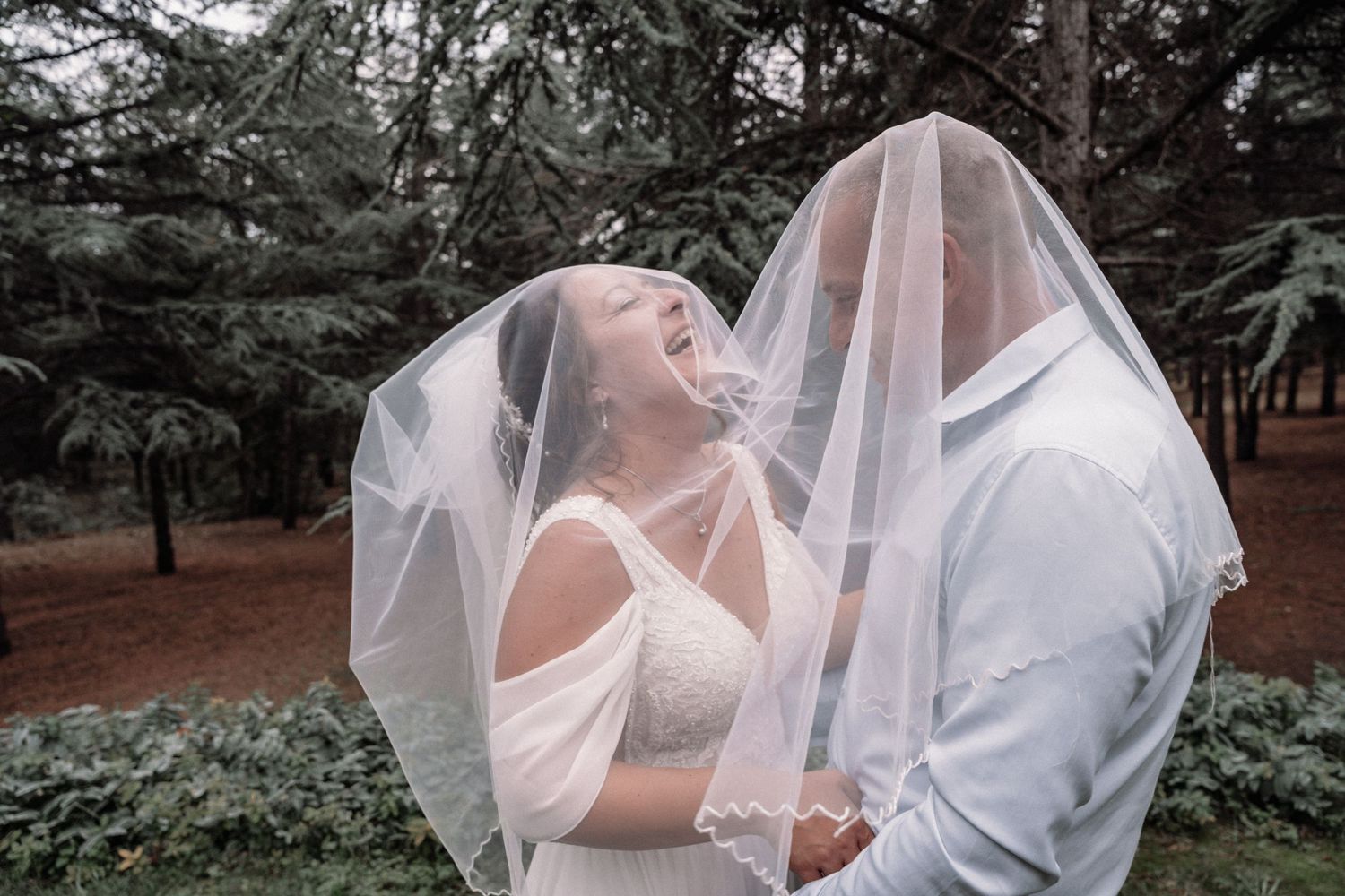 Playful wedding couple share romantic moment under flowing veil in forest setting.