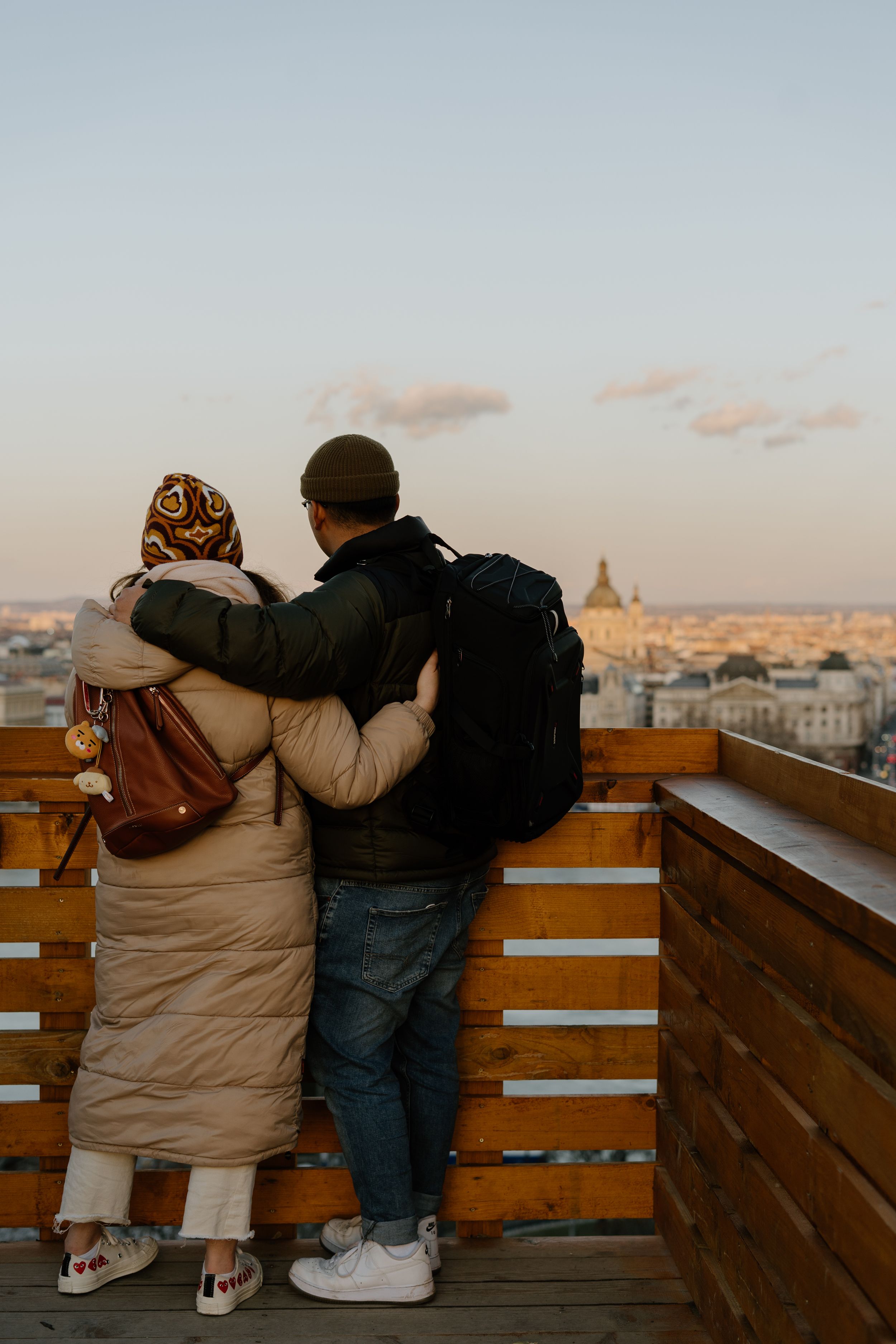 A couple stands on a wooden viewing platform overlooking a cityscape at sunset, embracing as they take in the view.