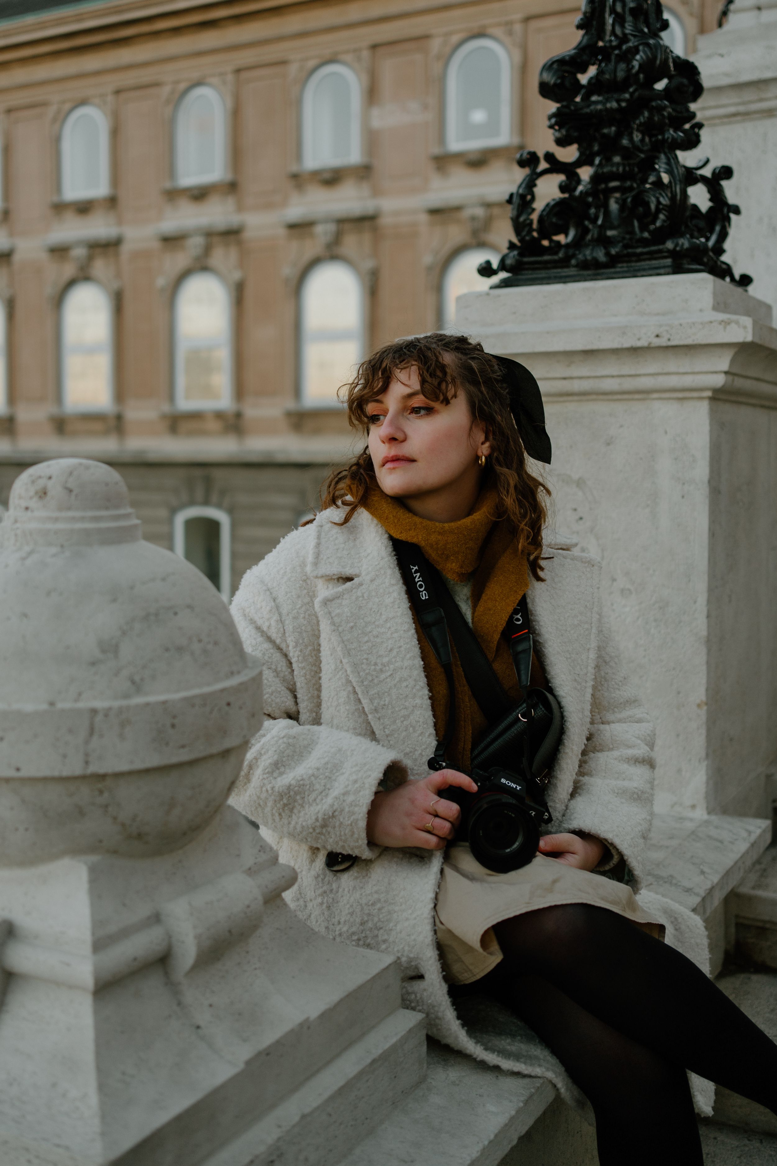 A photographer in a cozy cream sherpa coat and orange turtleneck sits on stone steps near a historic building exterior.