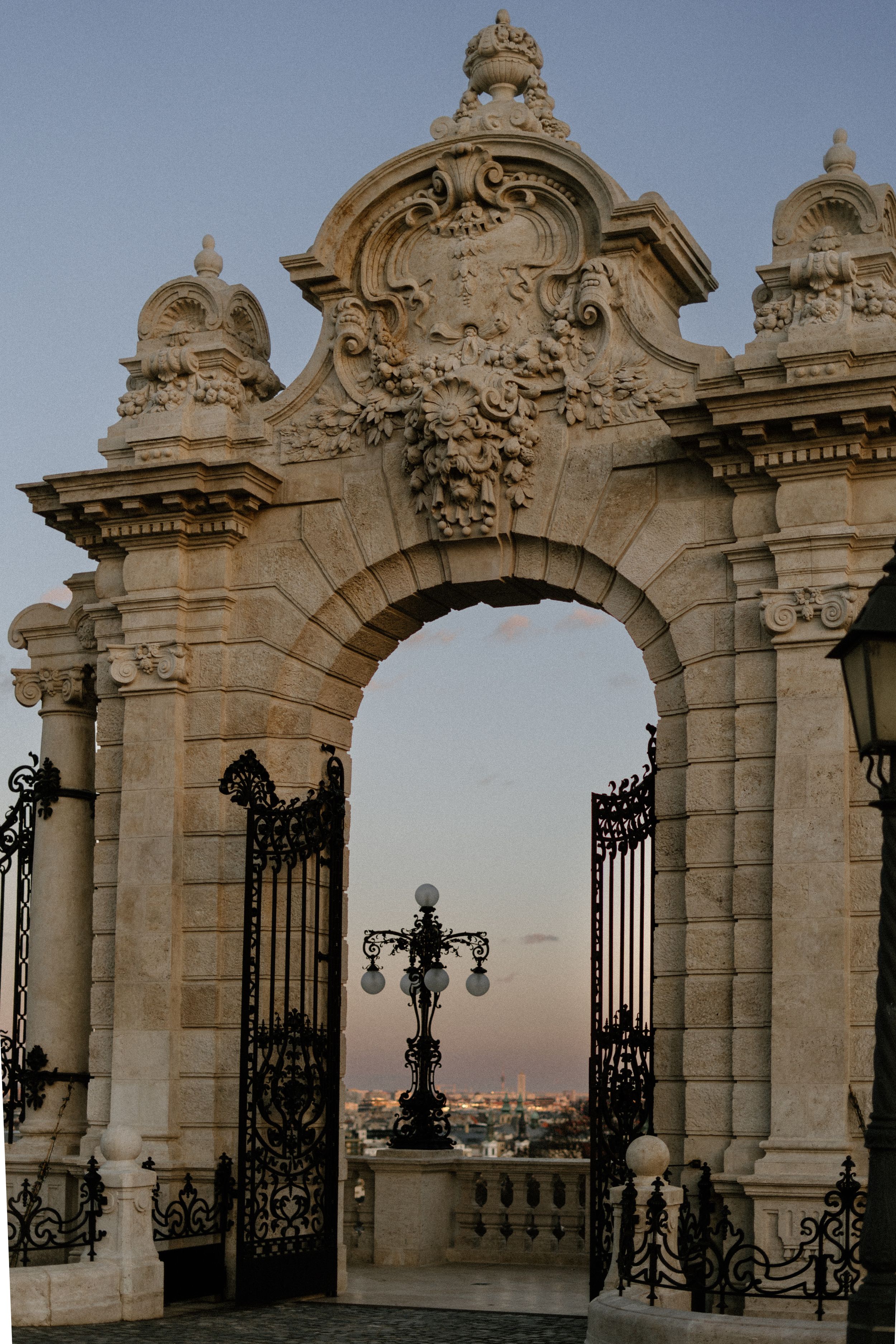 Ornate baroque stone archway with wrought iron gates overlooks Budapest cityscape at sunset from Buda Castle.
