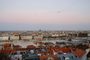 Panoramic cityscape of Budapest at sunset showing the Danube River and iconic red rooftops against a pastel sky.