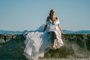 Elegant white wedding dress flowing in the breeze as woman sits on stone wall against bright blue sky and mountains. Nikola vjenčanje u ptuj
