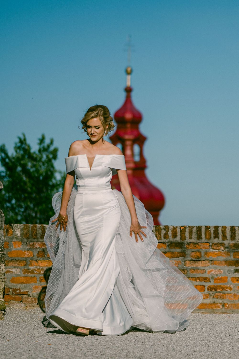 Elegant wedding dress with off-shoulder design and flowing train against brick wall with red church spire in background.