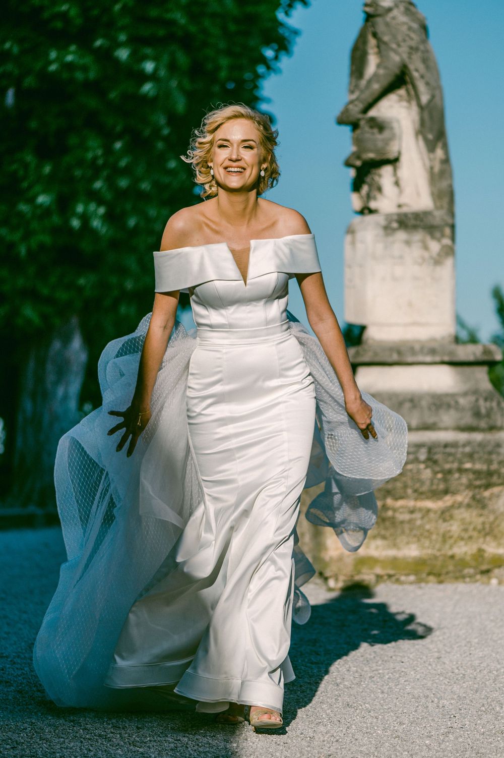 A woman in an elegant off-shoulder white wedding gown runs joyfully on a path with a statue visible in the background.
