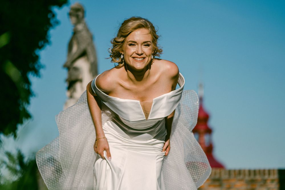 A person in a white strapless wedding dress poses joyfully in an outdoor garden setting with a statue in the background.