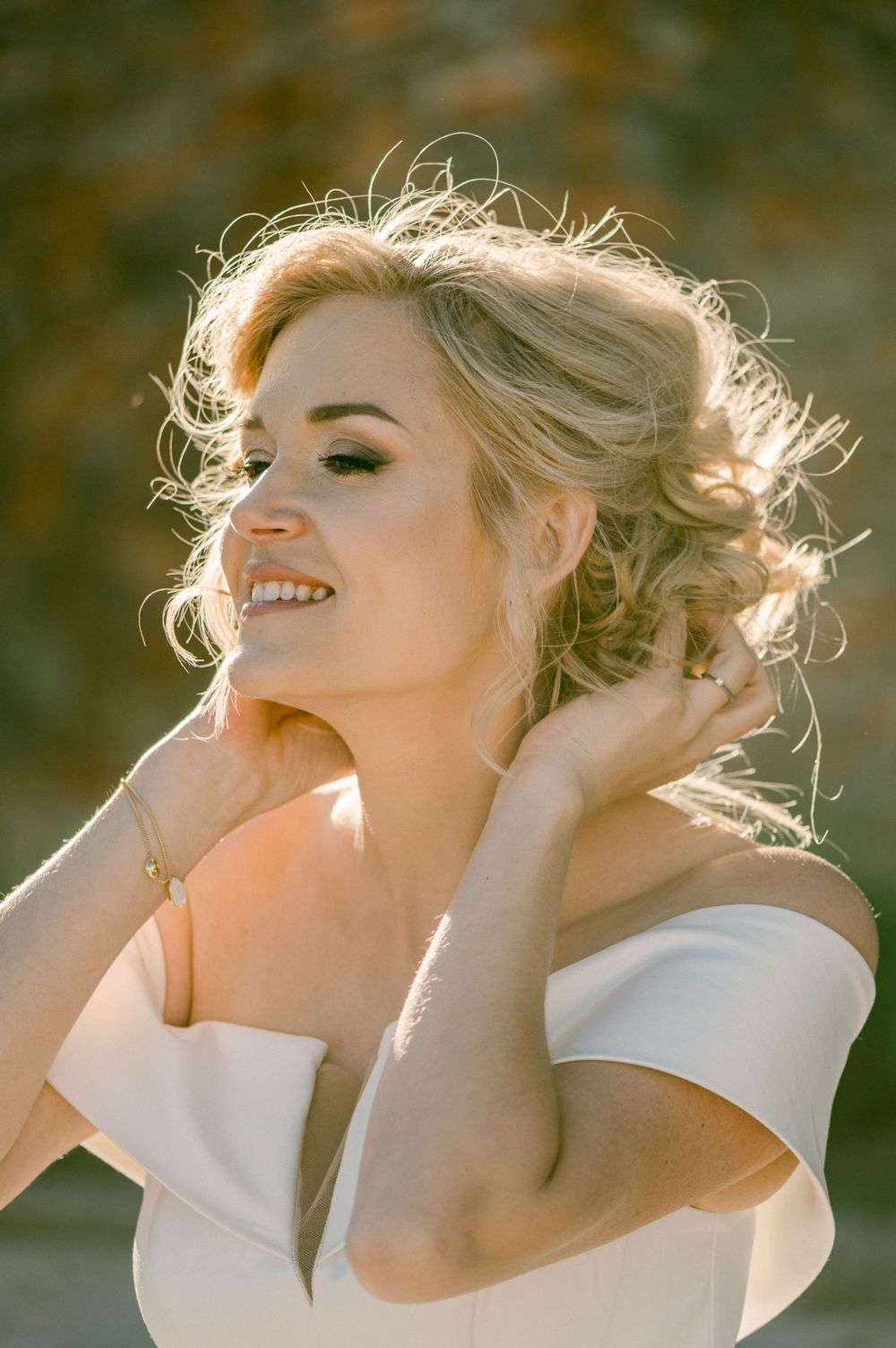 Portrait of a smiling person in sunlight wearing an elegant white off-shoulder dress, with backlit blonde hair.