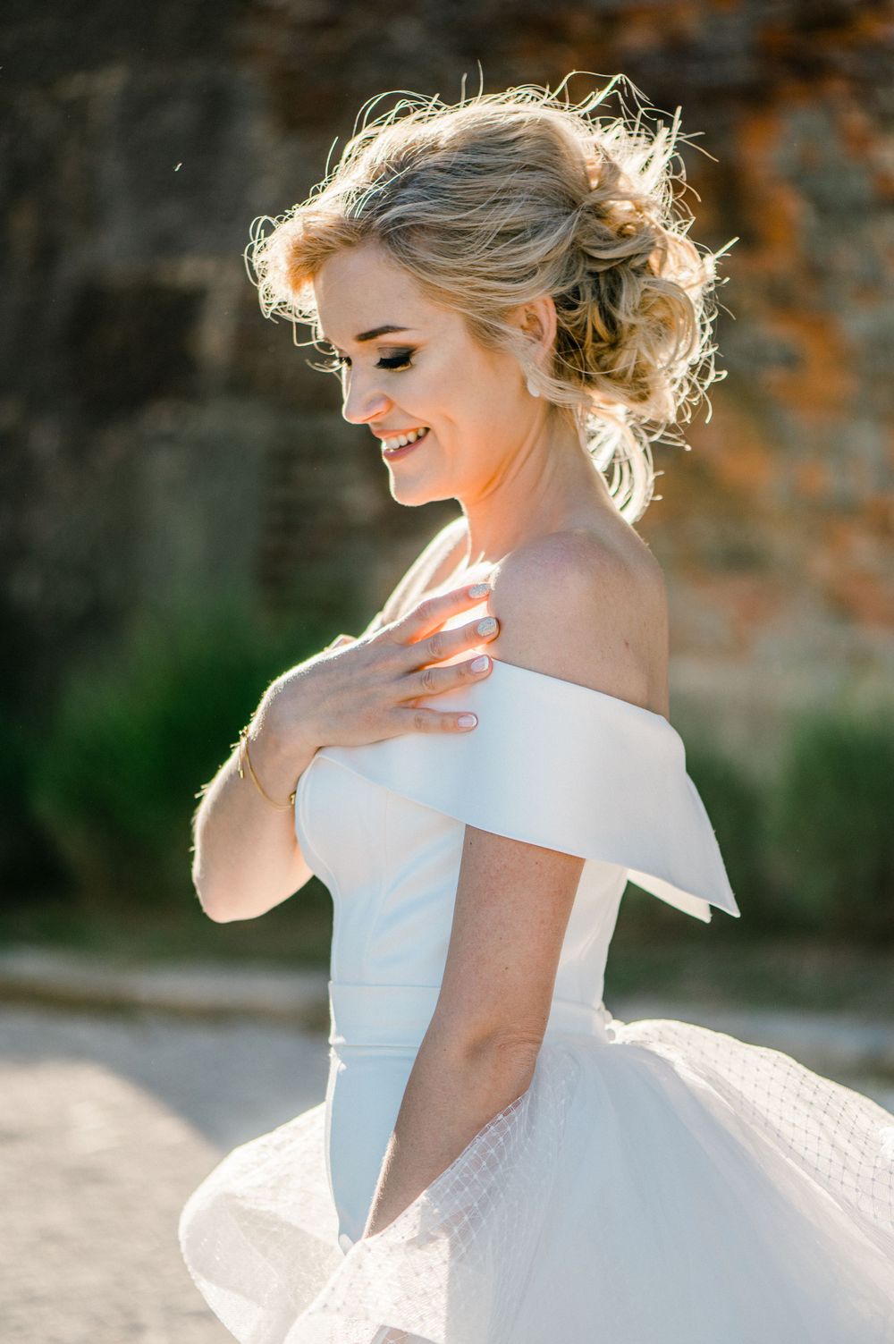 Ethereal bridal portrait in off-shoulder white gown with sunlit hair and natural outdoor backdrop.