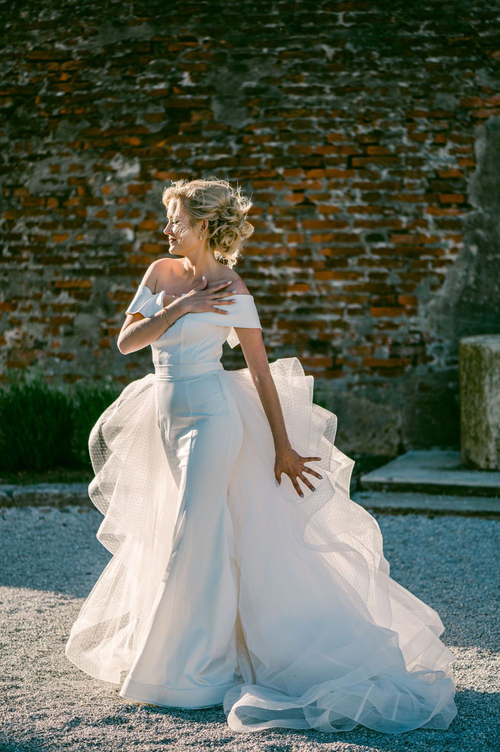 A bride in an elegant white off-shoulder wedding gown with flowing ruffles poses against a rustic brick wall background.