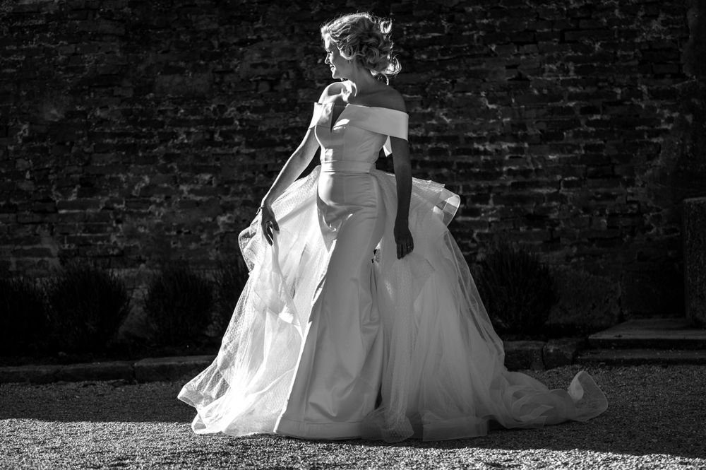 Black and white photo showing billowing white wedding dress fabric flowing in the breeze against a dark brick wall.