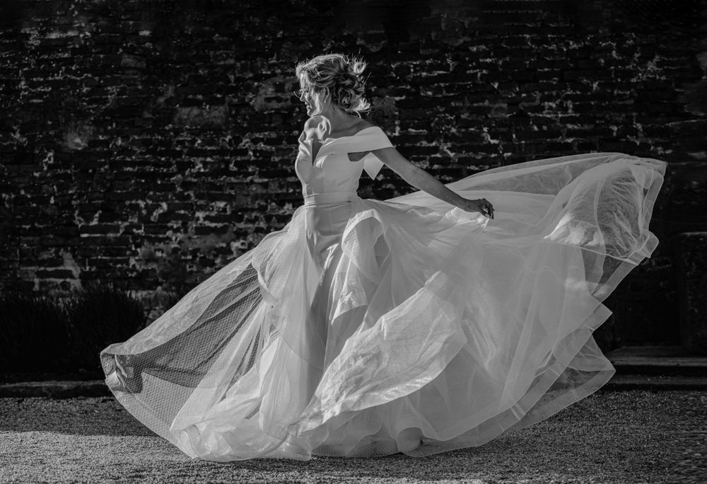 A flowing white wedding dress billows dramatically in the breeze against a dark brick wall background in black and white.