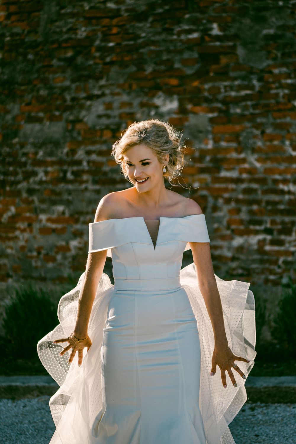 Elegant off-shoulder white wedding dress with fitted silhouette photographed against rustic brick wall in golden sunlight.