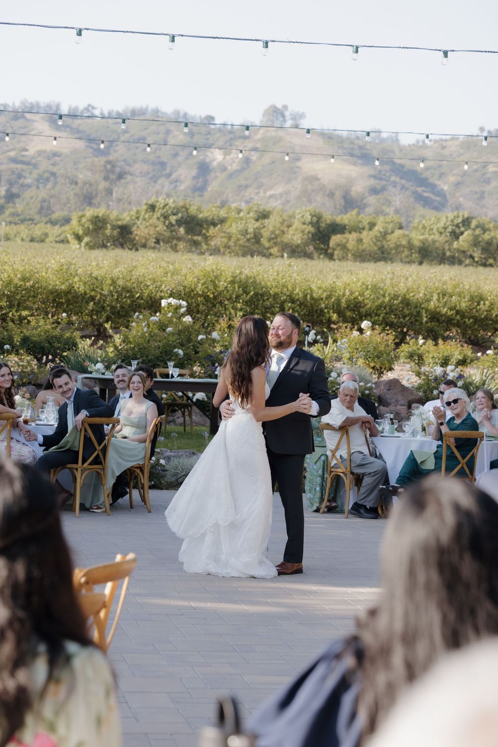 Bride and groom share their first dance outdoors at a vineyard wedding reception with guests seated around them.