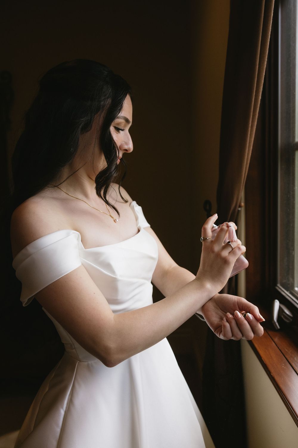 Bride in off-shoulder white gown applying perfume by a window, soft natural light highlighting her elegant look.