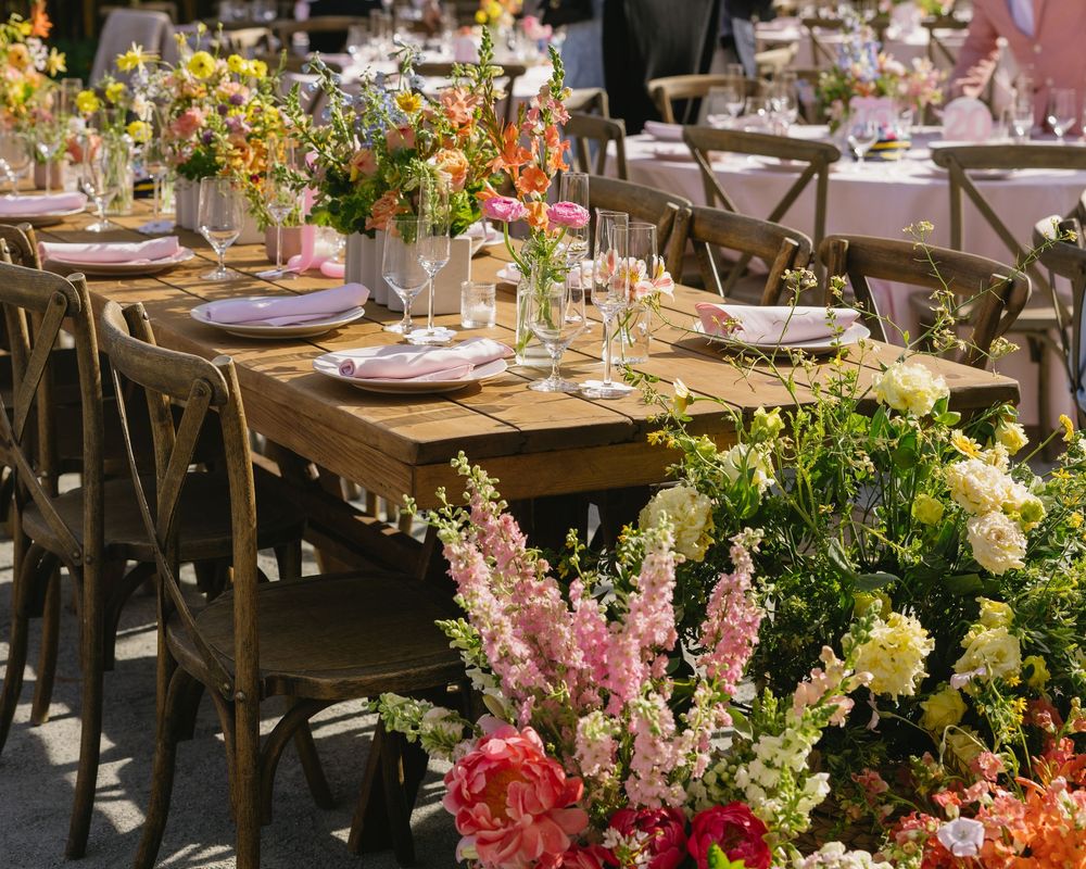 Elegant outdoor wedding reception with wooden farm tables adorned with colorful floral arrangements and pink place settings.