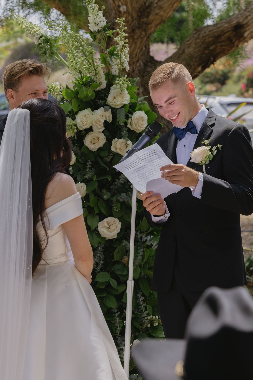 Groom smiling while reading wedding vows to bride at outdoor ceremony with white floral arch backdrop.