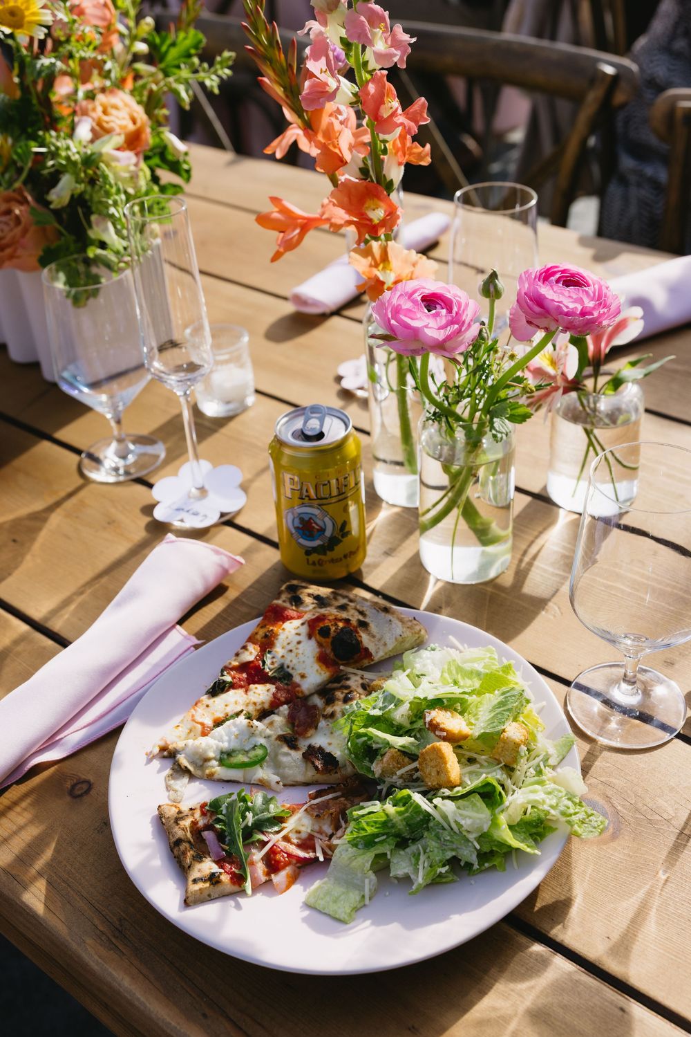 Outdoor dining table with pizza, Caesar salad, canned drink, wine glasses, and colorful floral arrangements in sunlight.