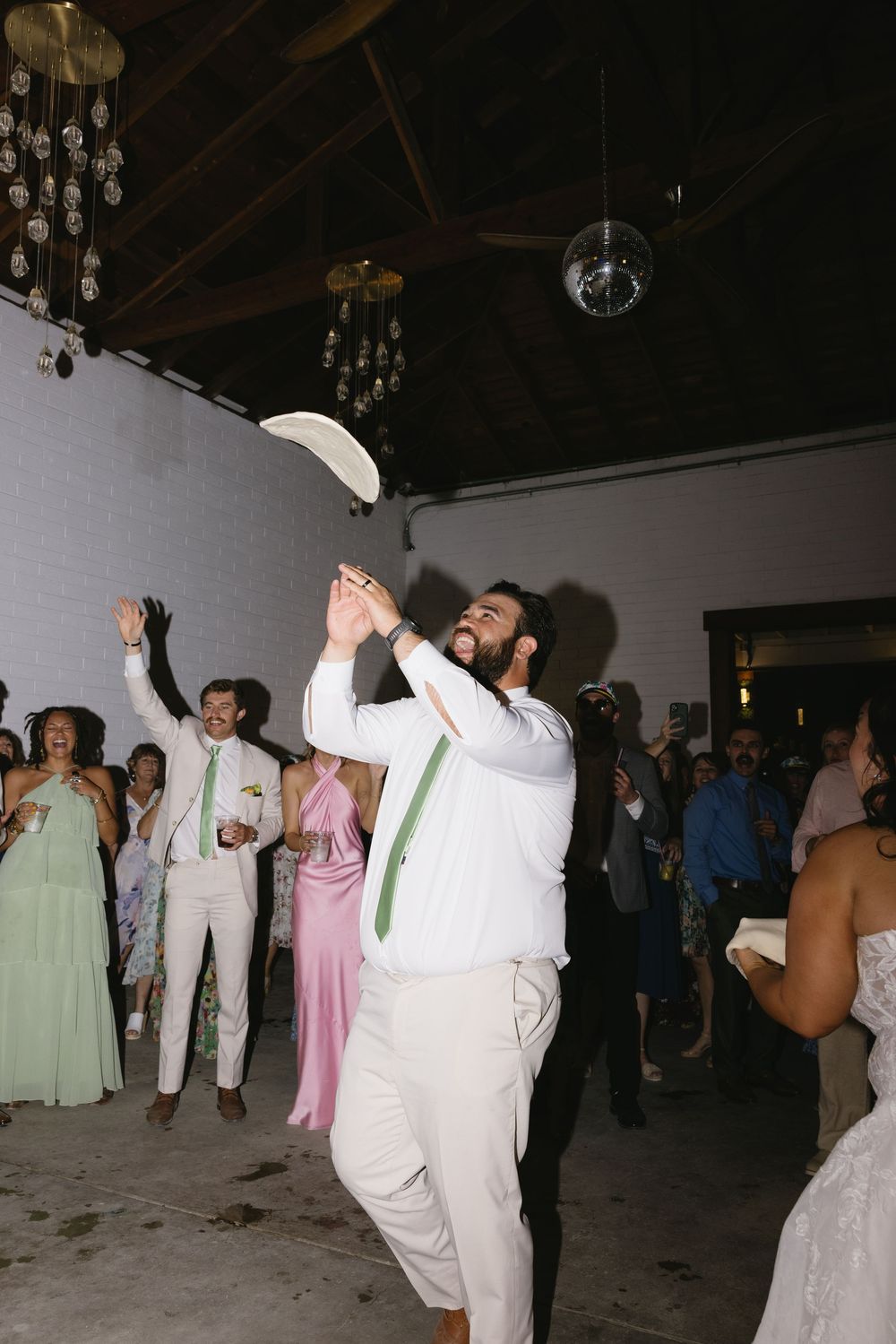 Groom in white shirt and green tie tossing his hat at a lively wedding reception with guests celebrating.