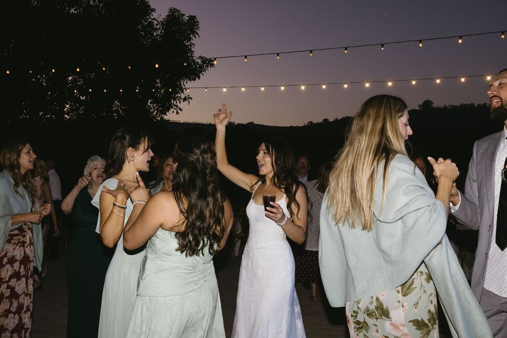 Guests dancing outdoors at a wedding reception under string lights at dusk, with bride in white dress.