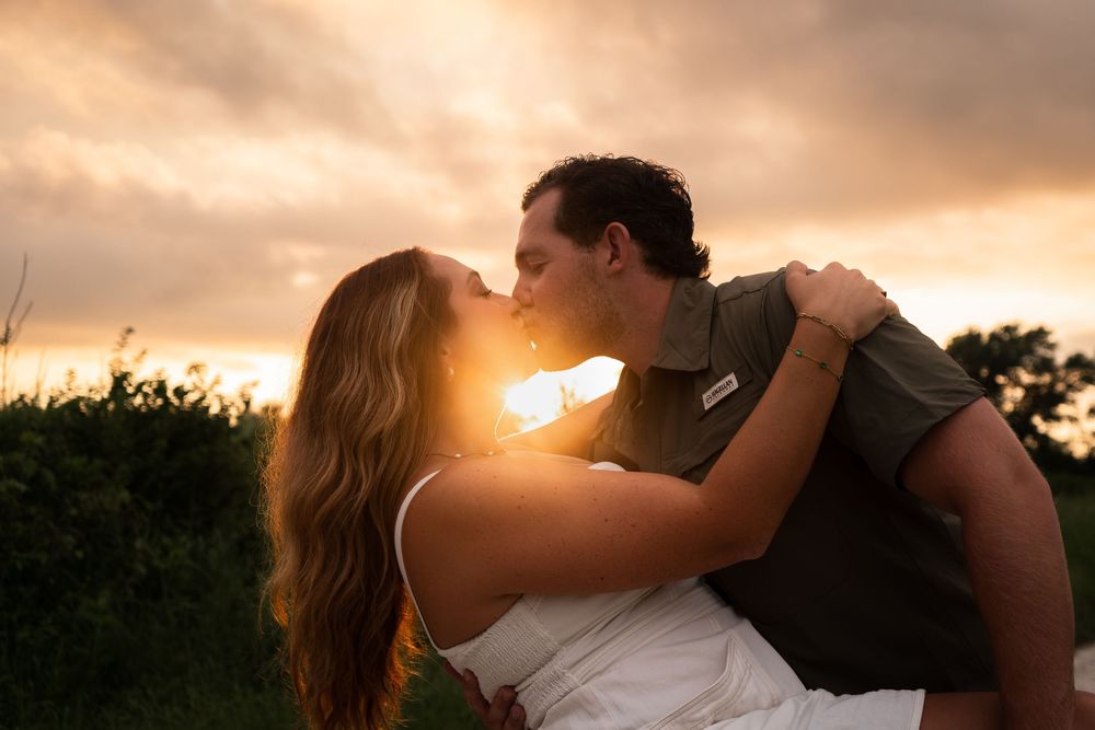 Silhouetted romantic couple sharing a passionate kiss against a warm sunset sky with golden light and trees.