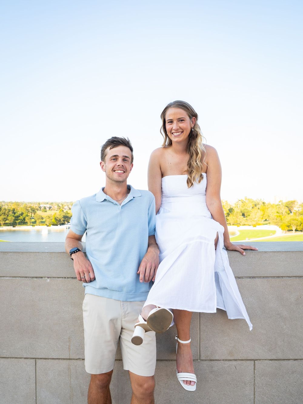 A couple in coordinating light blue and white outfits poses casually against a concrete wall with a sunny field behind them.